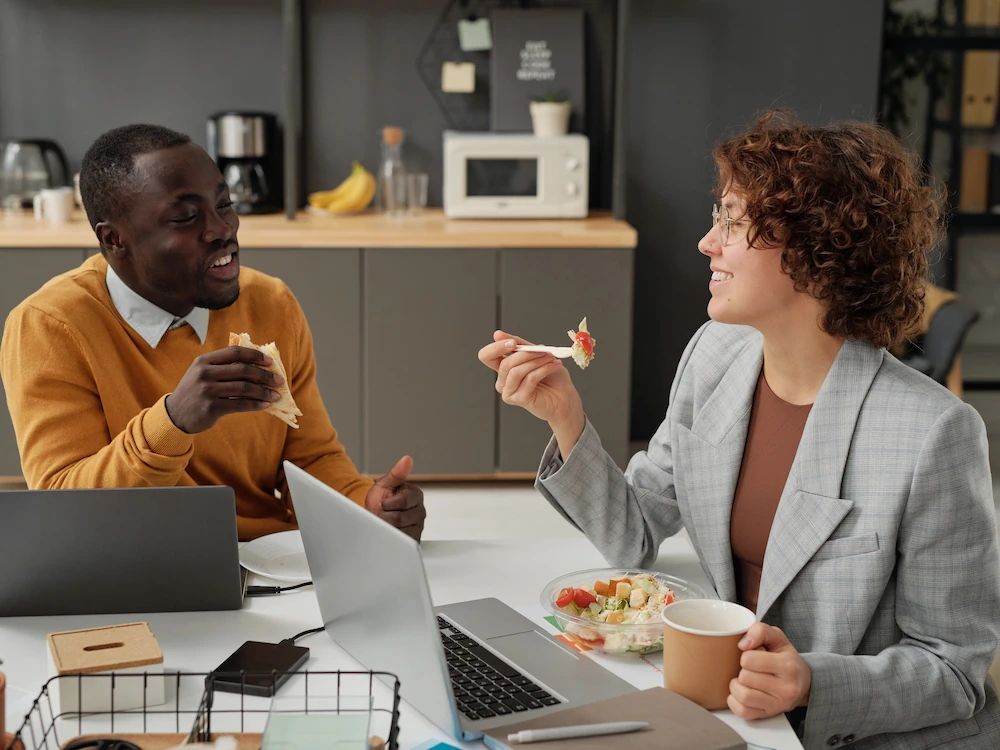 Man and woman having lunch in office setting while working on laptops