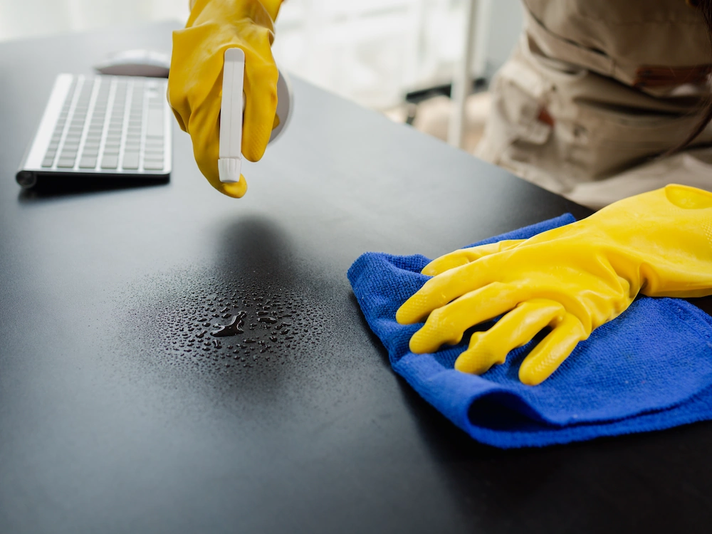 Janitorial worker with gloved hands spraying desk surface, wiping with microfiber cloth